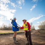 Two individuals in hardhats and high visibility outerwear shake hands on a worksite.