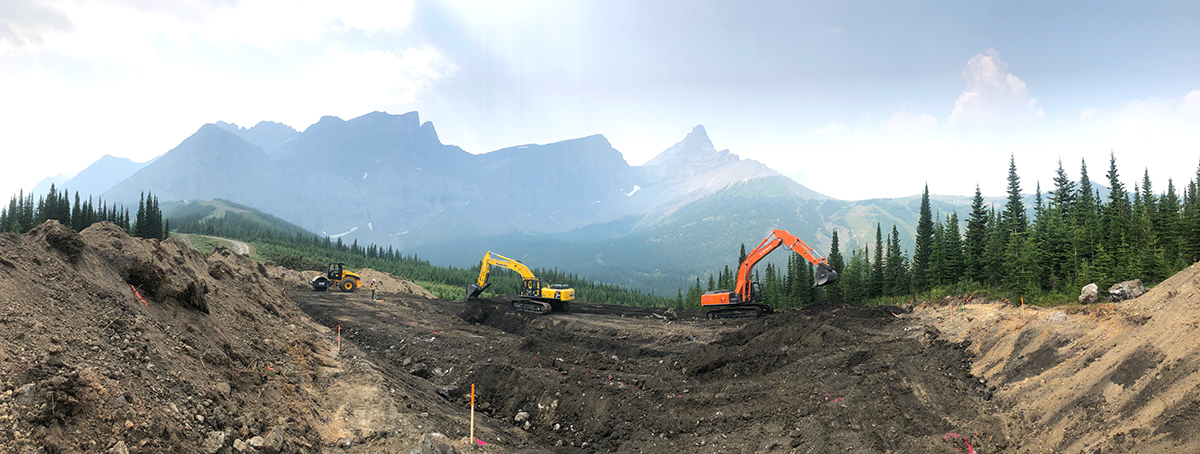 Fortress Mountain 1_02 Two excavators working in a cleared mountain area surrounded by forest, with rugged peaks in the background.