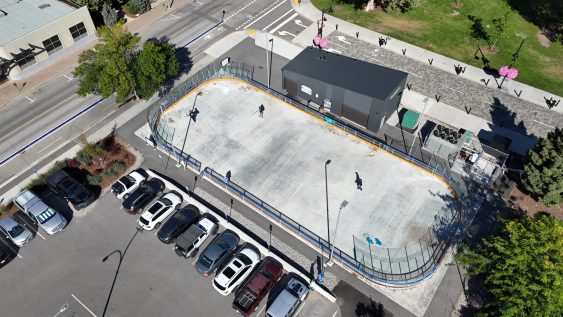 Aerial image of an outdoor rink in the summer season. Three street hockey players are visible in the rink. The rink is bordered by a scattering of buildings, and parked cars in a parking lot.