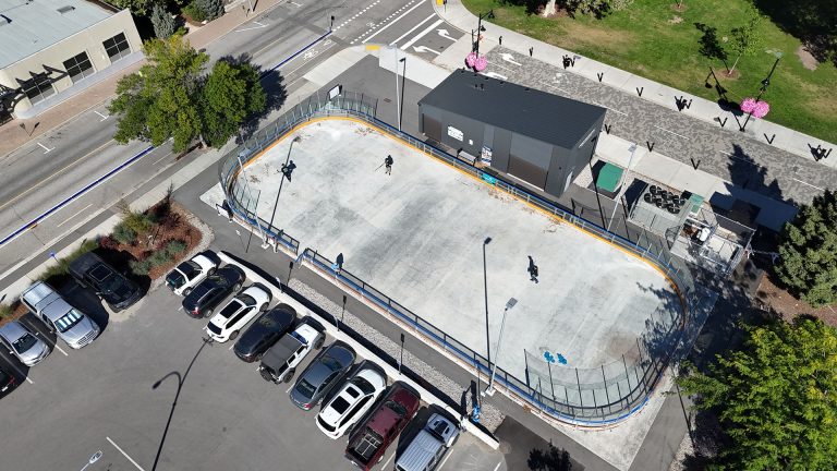 Aerial image of an outdoor rink in the summer season. Three street hockey players are visible in the rink. The rink is bordered by a scattering of buildings, and parked cars in a parking lot.