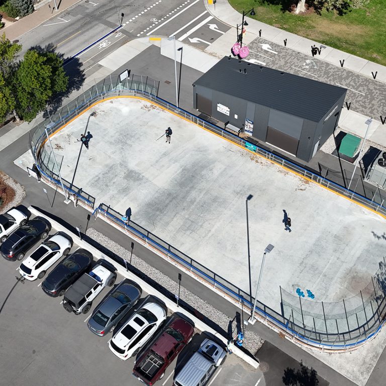 Aerial image of an outdoor rink in the summer season. Three street hockey players are visible in the rink. The rink is bordered by a scattering of buildings, and parked cars in a parking lot.