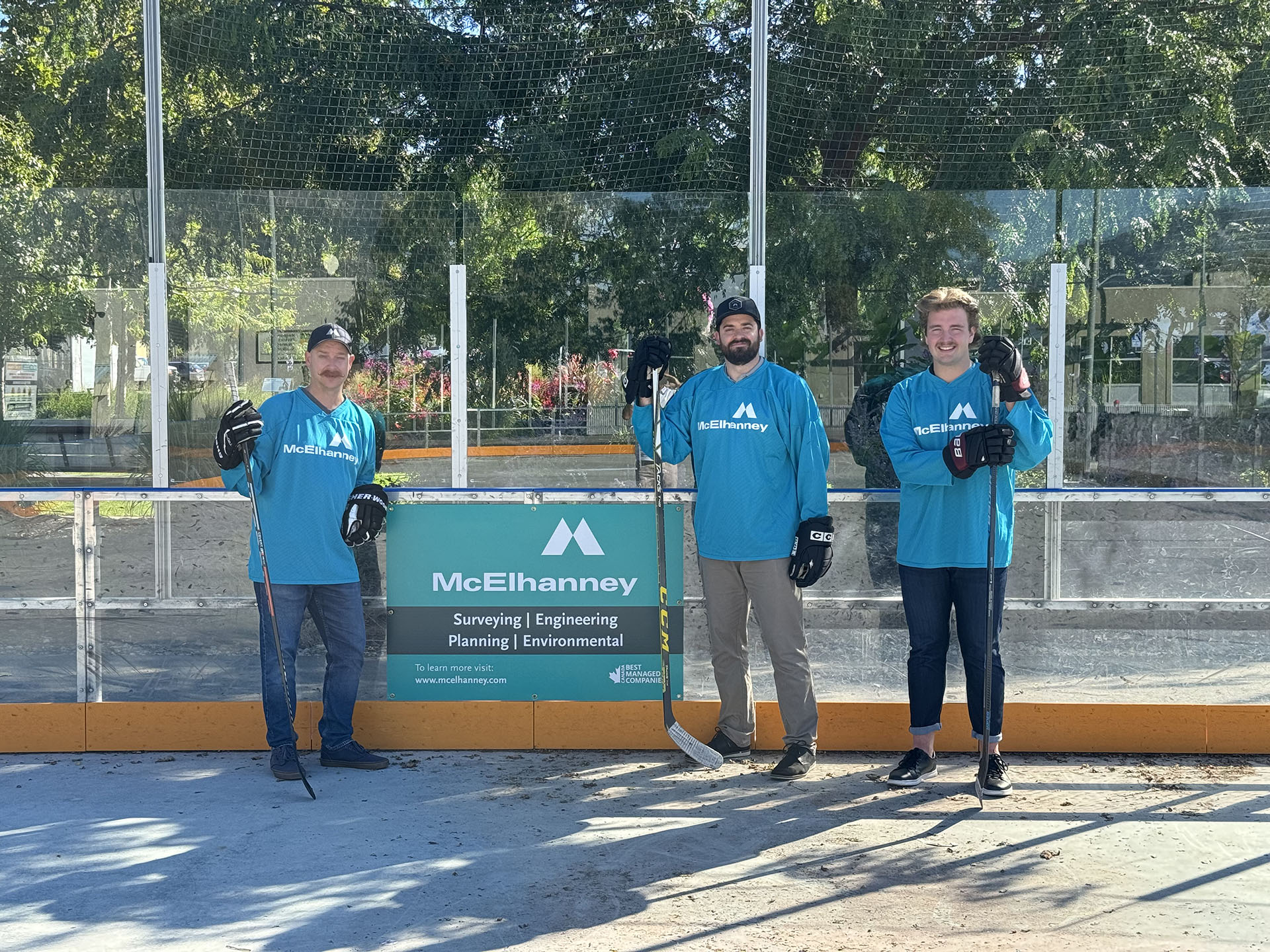 Penticton Outdoor Rink & McElhanney Hockey Team Three individuals with street hockey equipment, including hockey gloves, sticks, and teal jerseys featuring McElhanney's logo, stand in an outdoor skating rink around a McElhanney promotional sign.
