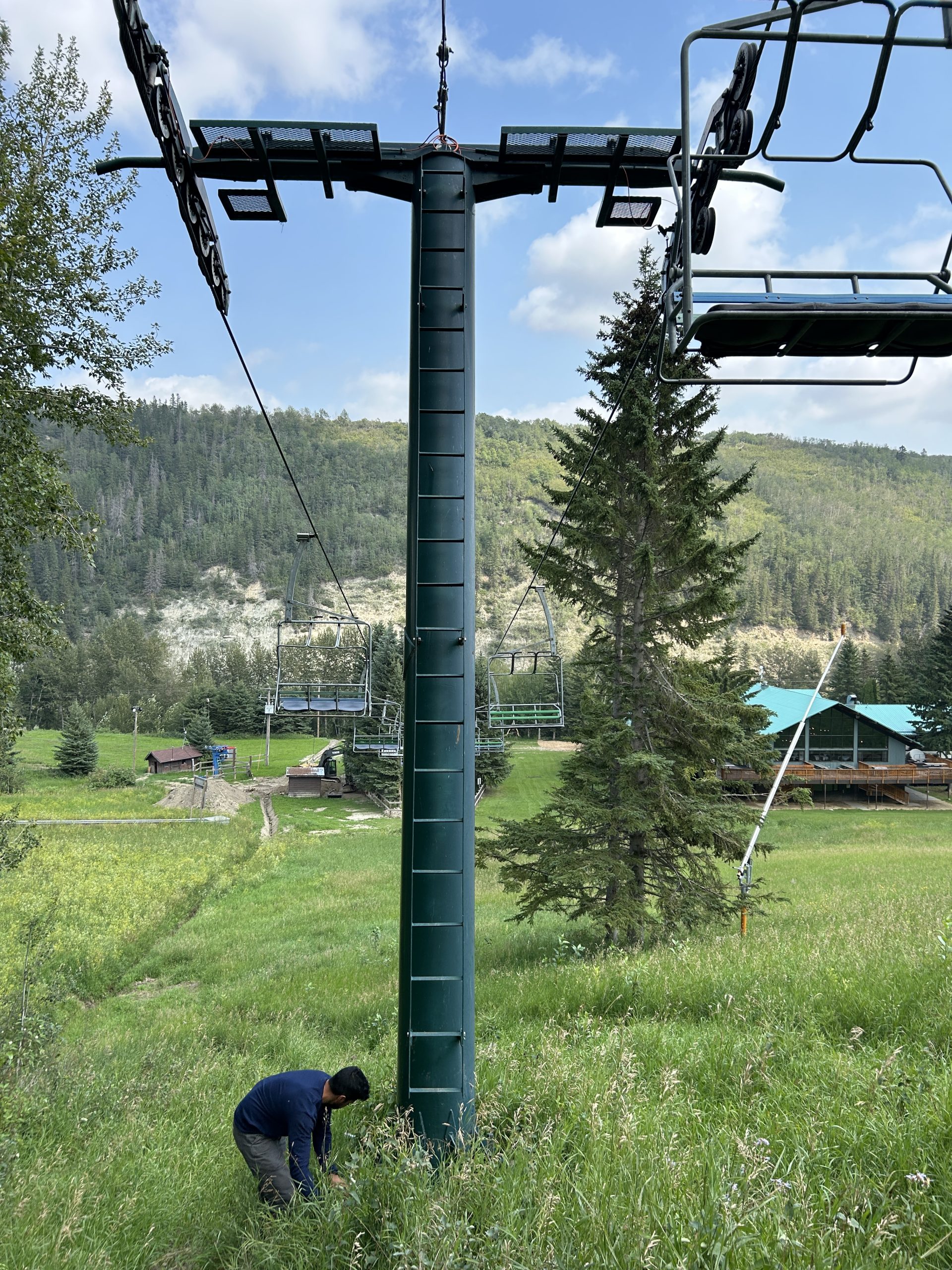 Canyon Ski Lift Upgrades A blue chairlift tower is in the foreground. The tower is surrounded by grassy slopes and there are hills in the background. A ski lodge is visible in the bottom, right-hand corner.