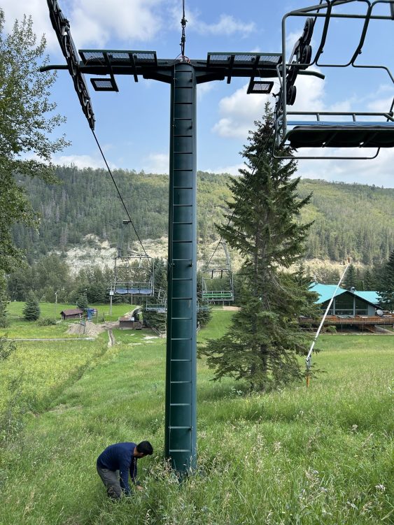A blue chairlift tower is in the foreground. The tower is surrounded by grassy slopes and there are hills in the background. A ski lodge is visible in the bottom, right-hand corner.
