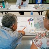 Two individuals evaluate and mark up drawings spread out on a table in front of them. Markers and pencils are scattered around.