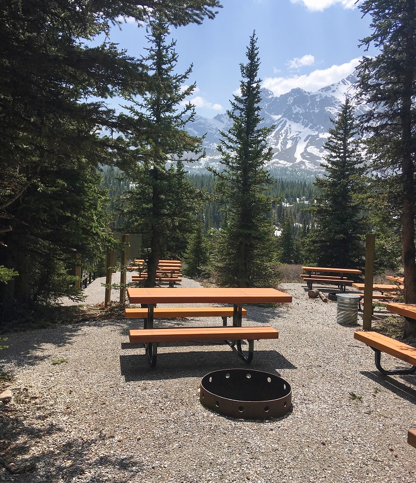 Fire pit and multiple picnic tables in a remote backcountry campground with a backdrop of mountains.