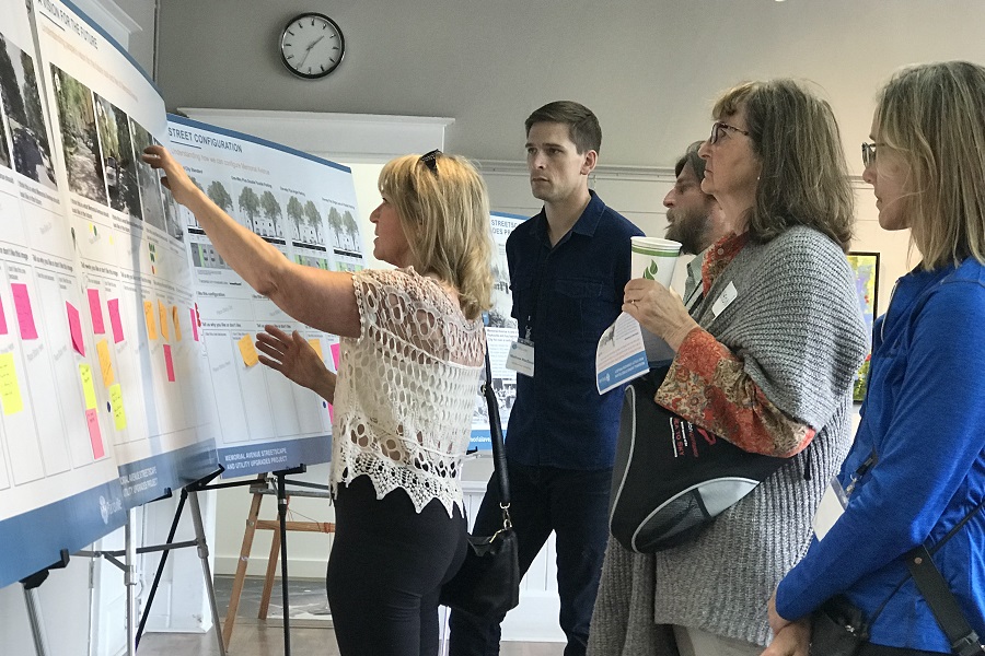 There is a series of easels displaying poster featuring project related content. A group of people is gathered around the posters. One onlooker is reaching towards a poster and making an observation about it.