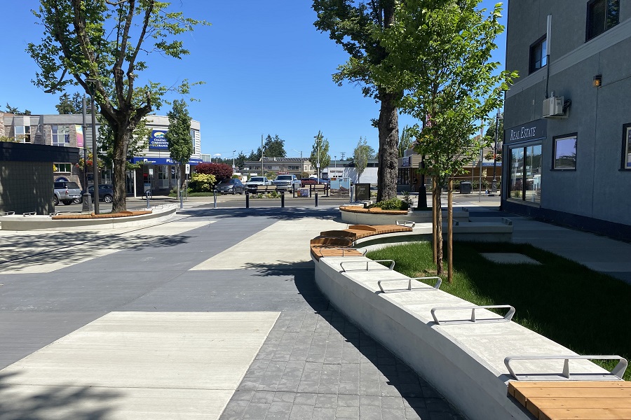 A boulevard of mixed paved surfaces expands outward towards a tree-lined parking lot. There is curving concrete bench lining the boulevard.
