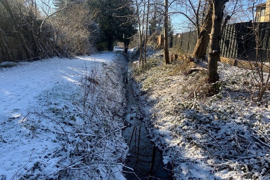 A shallow, snow-covered ditch is filled with a low level of water.
