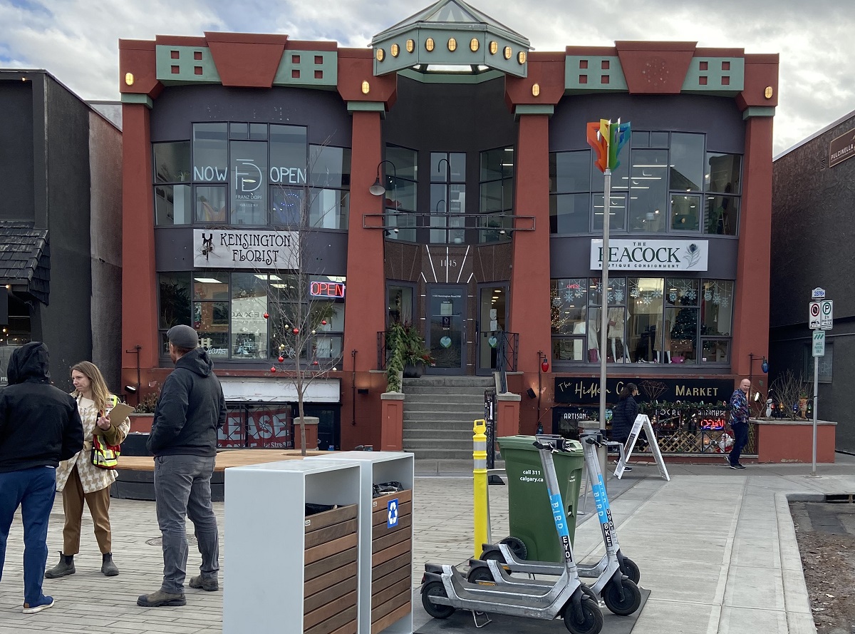 A two-story building with brick and window exterior is in the background. The plaza in the foreground has large paving blocks, scooter parking, and a tree and bench. There are three people conversing towards the side of the image.