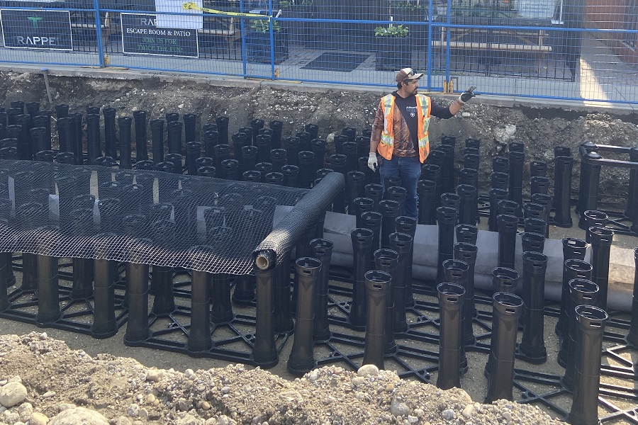 A construction worker stands a few feet below grade, surrounded by vertical, black cylinders.
