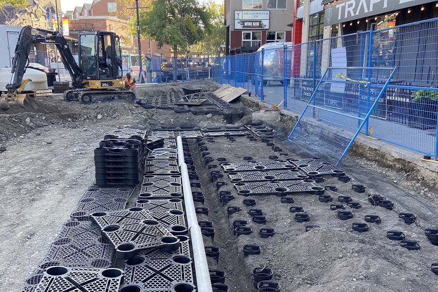 Soil has been backfilled around vertical, black cylinders that reach a few feet below grade. Blue construction fencing separates the work area from the adjoining shops. An excavator is parked in the background.