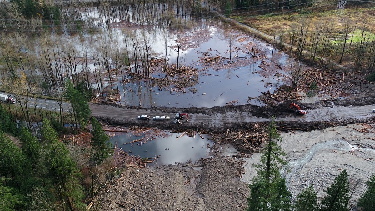 A highway is in mid-repair. A gravel section suffices for the roadbed where the pavement has been washed away by heavy rainfall. Flooded landscape is visible on either side of the damaged roadway.