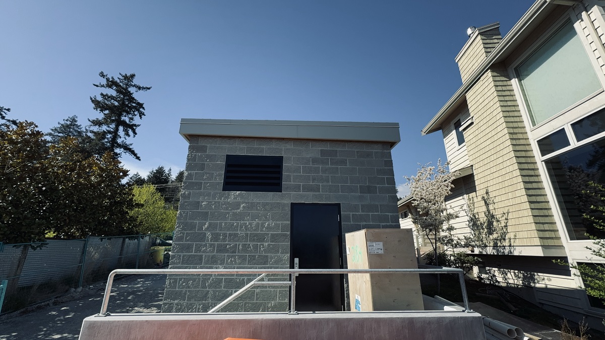 A narrow, single-story concrete structure is in the centre of the photo. The side of a house is visible on the right, a few metres away from the concrete structure. To the left of the concrete structure is a narrow laneway that is bordered immediately by a fence line and trees.