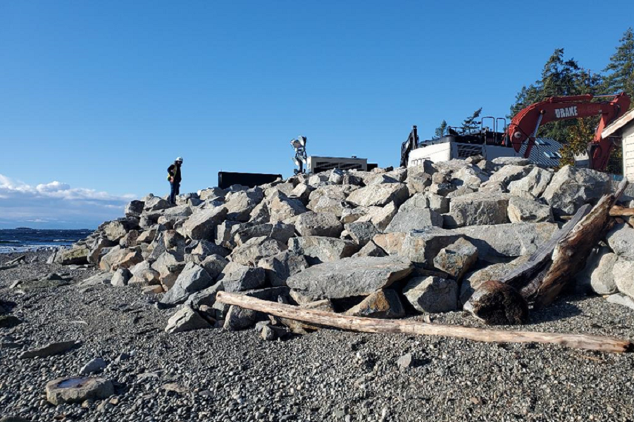 Strategically placed boulders create a sloping fortification wall on a rocky beach. Two construction workers and an excavator are visible at the top of the wall.