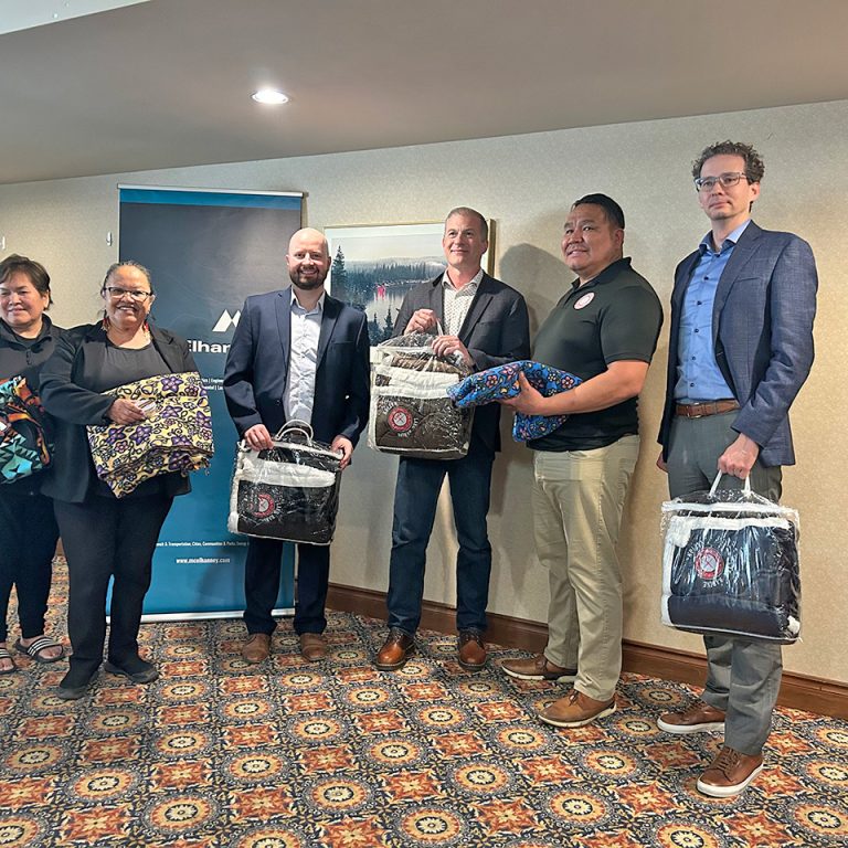 Three Indigenous elders and three McElhanney team members pose for a photograph in a meeting room. All six individuals are holding gifted blankets.
