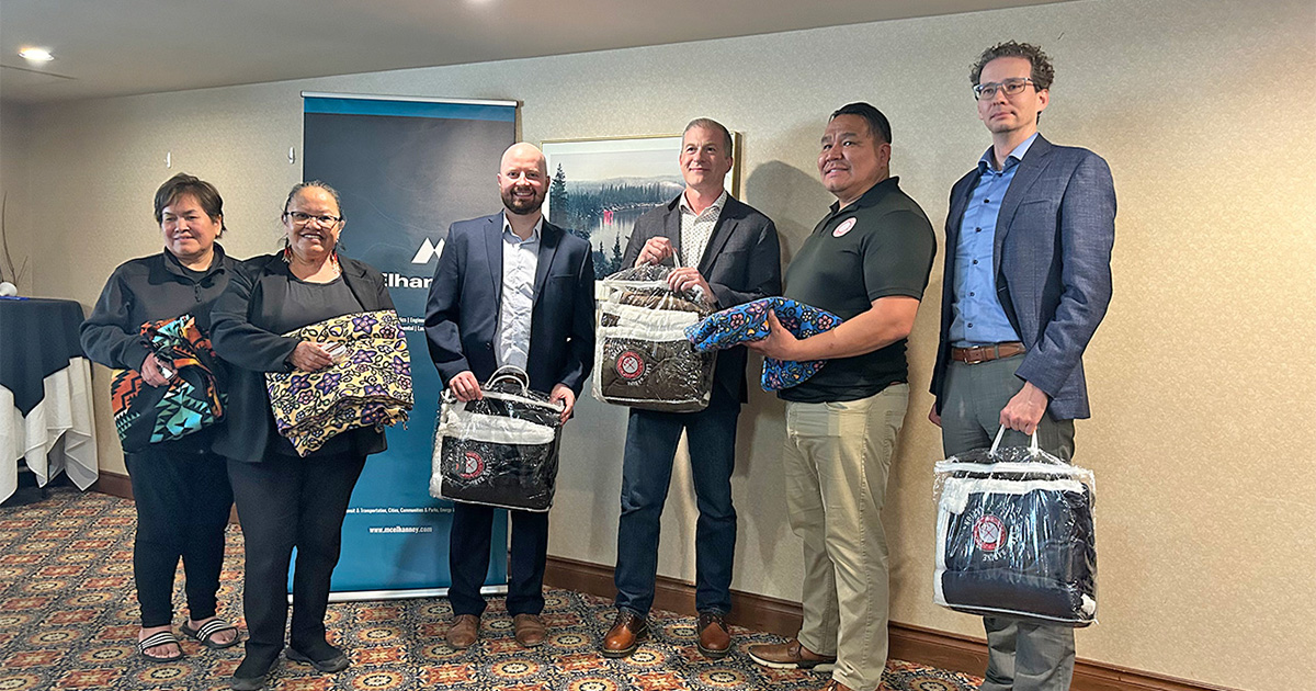 Three Indigenous elders and three McElhanney team members pose for a photograph in a meeting room. All six individuals are holding gifted blankets.