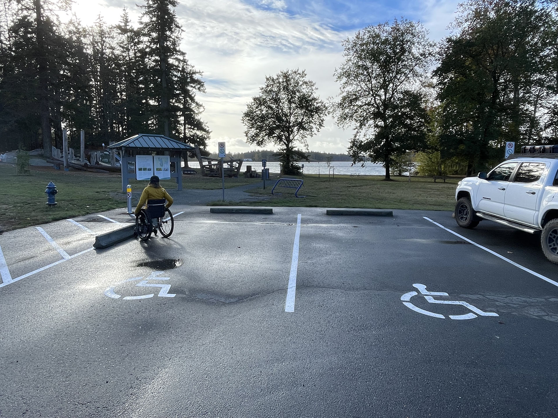 A person in a wheelchair has their back to camera and is in a parking lot, facing an interpretive sign at an outdoor recreational venue. There are two accessible parking spaces visible in the parking lot. There are trees in the background, and a body of water on the horizon.