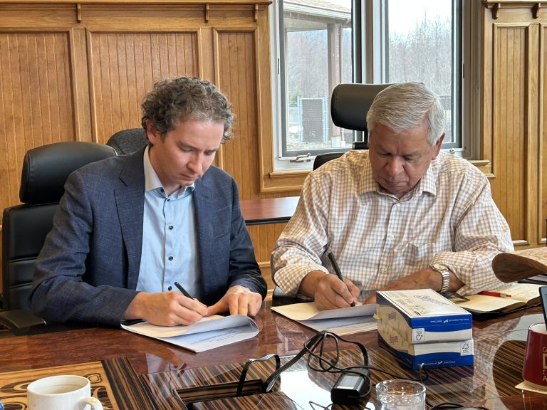 Two individuals in a corporate board room signing paper documents with pens.