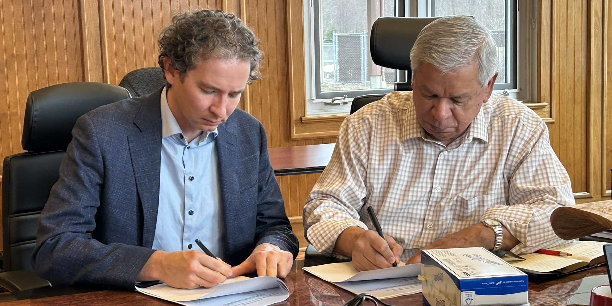 Two individuals in a corporate board room signing paper documents with pens.