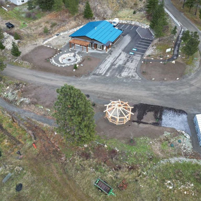 Skuppah’s Food Security Centre Aerial image showing green-roofed building on one side of a dirt road. A greenhouse and timber frame gazebo-style structure are visible across the road.