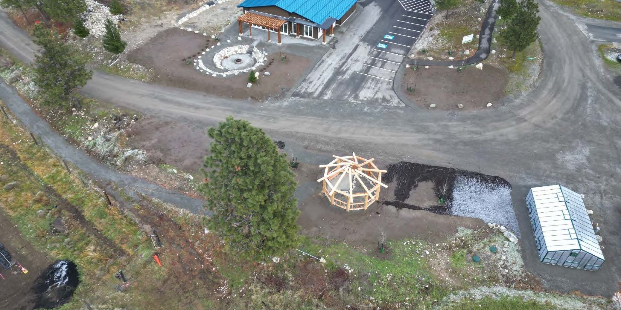 Aerial image showing green-roofed building on one side of a dirt road. A greenhouse and timber frame gazebo-style structure are visible across the road.