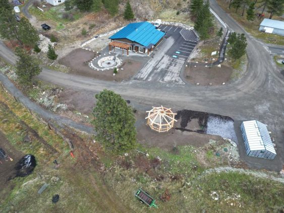 Aerial image showing green-roofed building on one side of a dirt road. A greenhouse and timber frame gazebo-style structure are visible across the road.