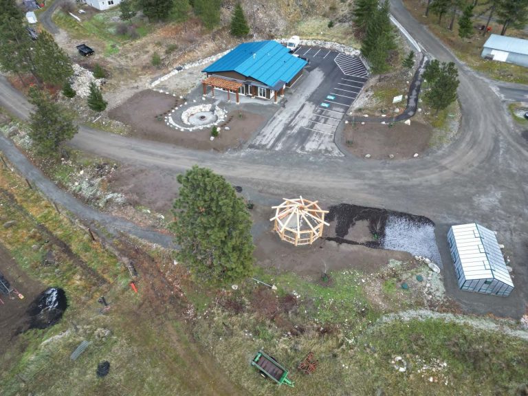 Aerial image showing green-roofed building on one side of a dirt road. A greenhouse and timber frame gazebo-style structure are visible across the road.