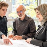 Three individuals of varying ages are gathered around, and discussing, a set of drawings.