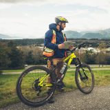 An individual in a high visibility vest and bike helmet stands alongside a bike on a gravel trail. The individual is holding a clipboard and visibly evaluating something off-camera.
