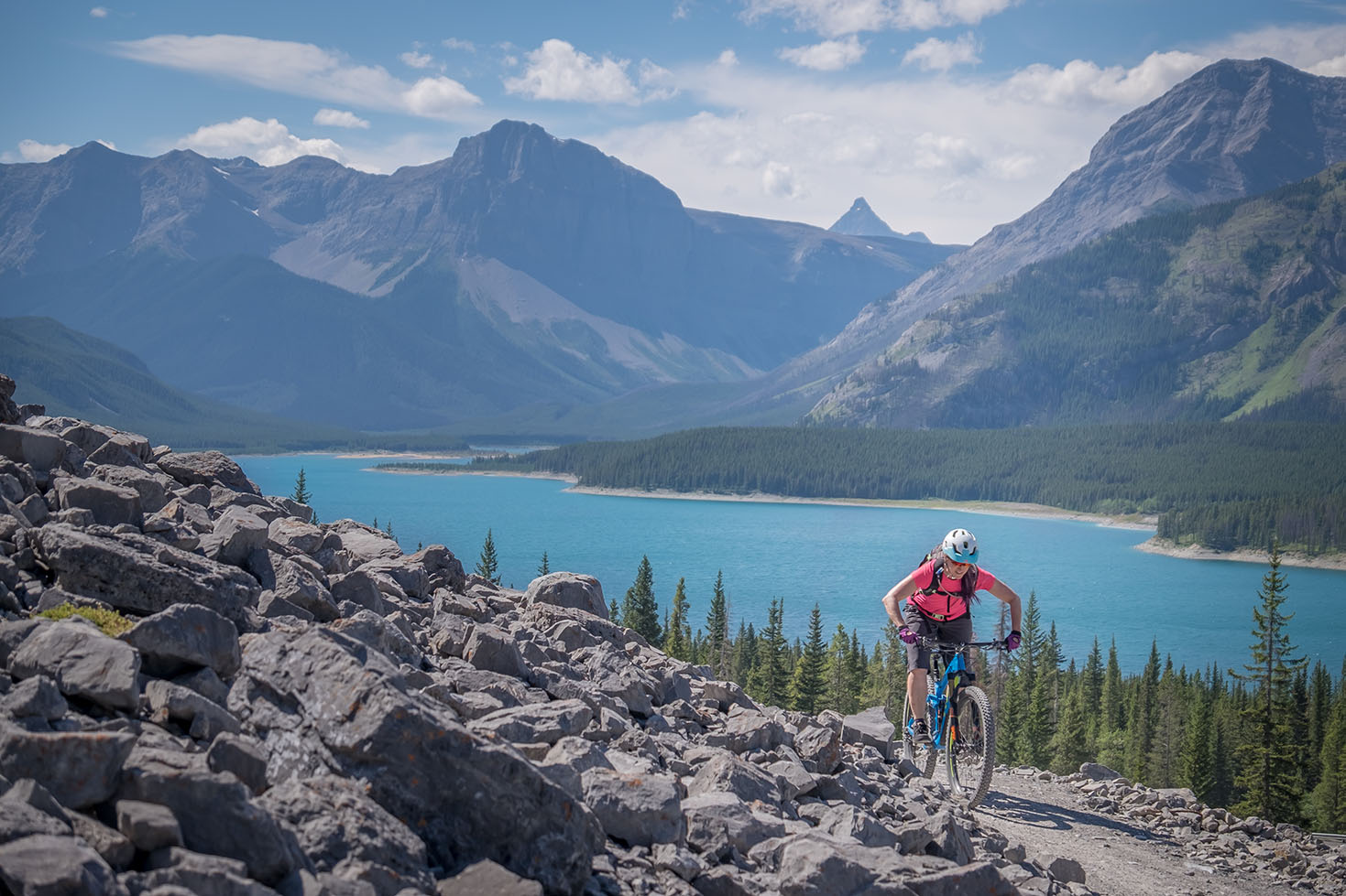A mountain biker rides up a rocky trail with a turquoise lake and dramatic mountain peaks in the background under a bright blue sky.
