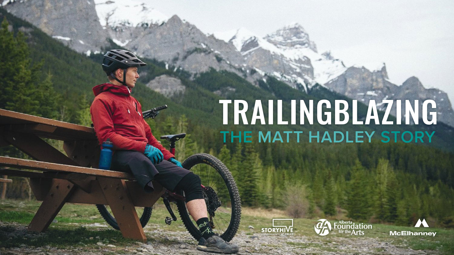 A man in mountain biking gear sits on a wooden picnic bench beside his bike, gazing at snow-capped mountains in the distance. Text reads “Trailblazing: The Matt Hadley Story,” with sponsor logos from STORYHIVE, Alberta Foundation for the Arts, and McElhanney.