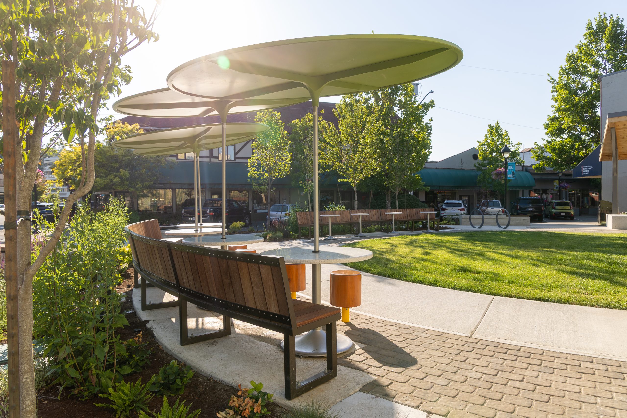 Benches and tables under large green shade structures in a landscaped urban park.