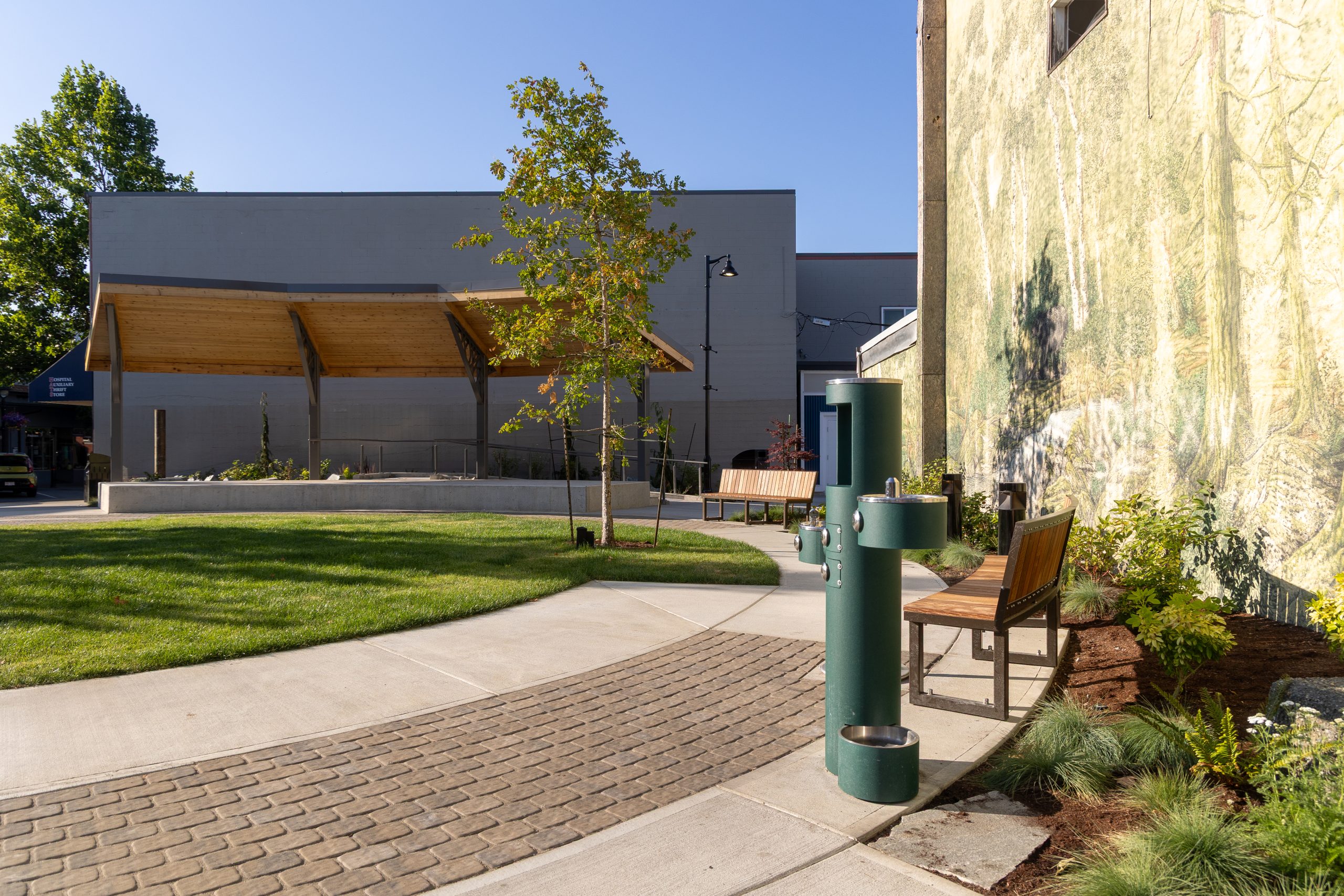 Park with open lawn, drinking fountain, benches, and a forest mural on a building wall.