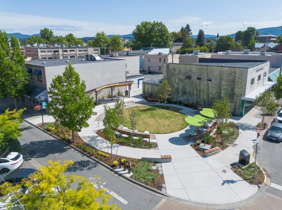 Aerial view of a downtown park with circular lawn, benches, trees, and a forest mural on a nearby building.