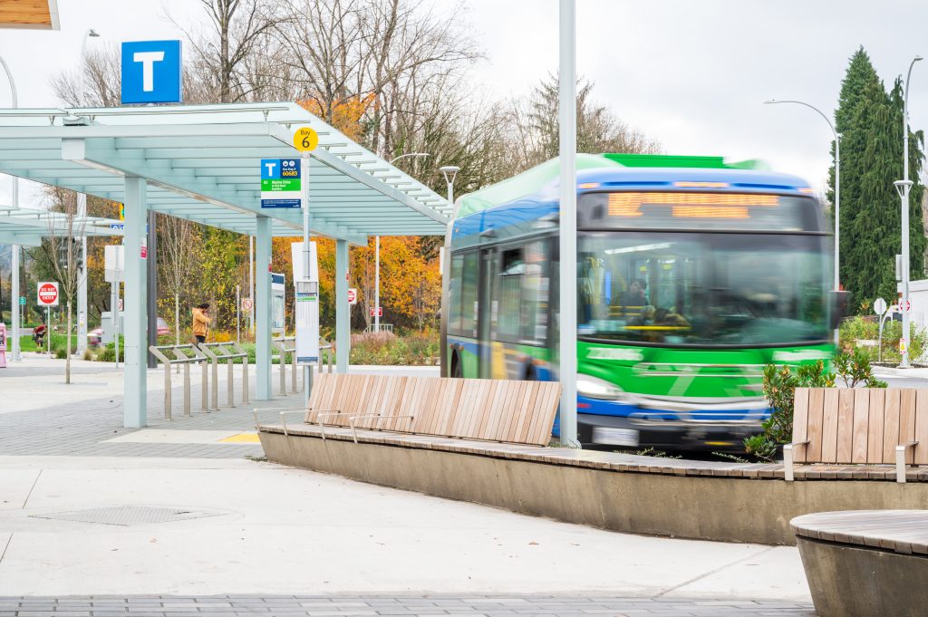 A green and blue bus in motion at the Phibbs Exchange Bus Station, with a covered waiting area, route signage, and autumn trees in the background.