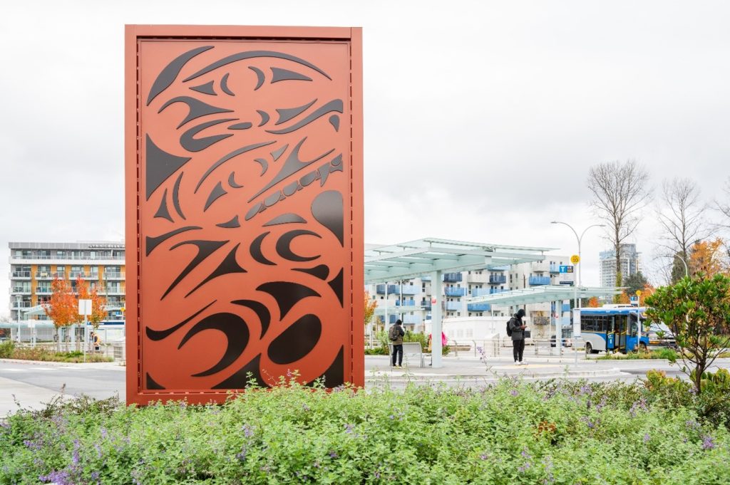 A close-up of an orange metal panel with black abstract designs beside a modern transit station with glass canopies and people waiting.