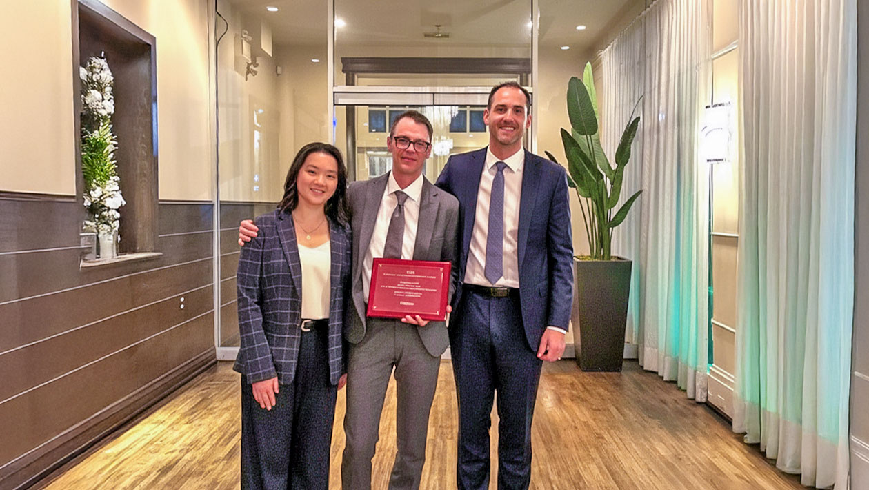 Three individuals in formal business attire stand in a modern hallway. The person in the center holds a red award plaque, suggesting a recognition moment at the 2025 CCE Award Gala.