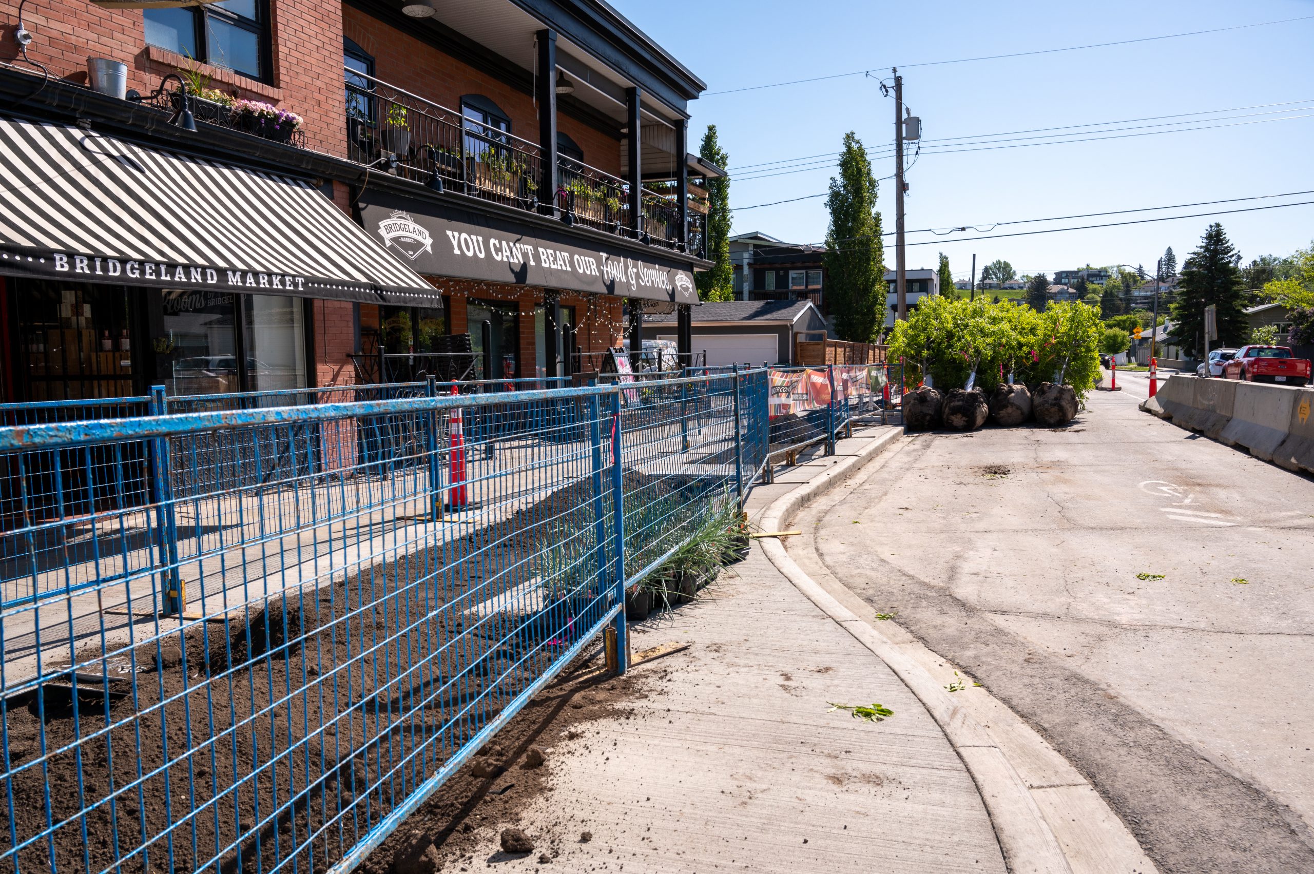 Streetscape construction outside Bridgeland Market, with fencing, soil work, and trees staged for planting along the sidewalk.