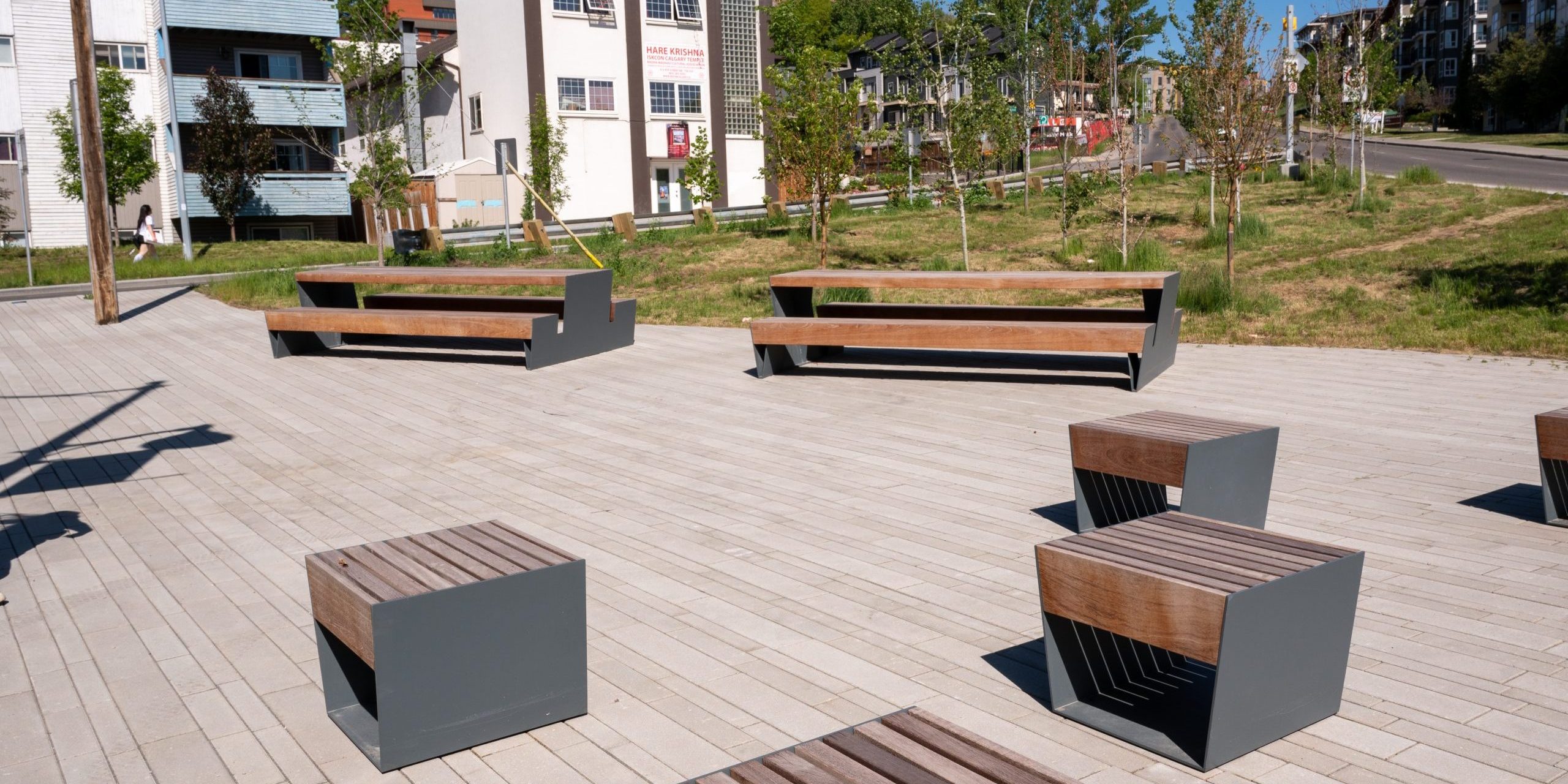 Modern outdoor plaza with wooden and metal benches and stools on paved stone, surrounded by new trees and residential buildings.