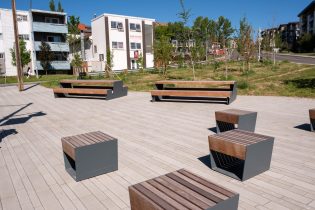 Modern outdoor plaza with wooden and metal benches and stools on paved stone, surrounded by new trees and residential buildings.