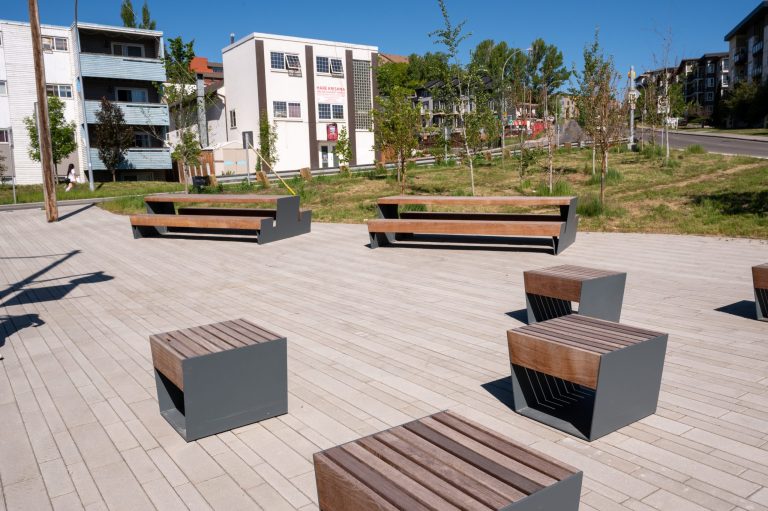 Modern outdoor plaza with wooden and metal benches and stools on paved stone, surrounded by new trees and residential buildings.