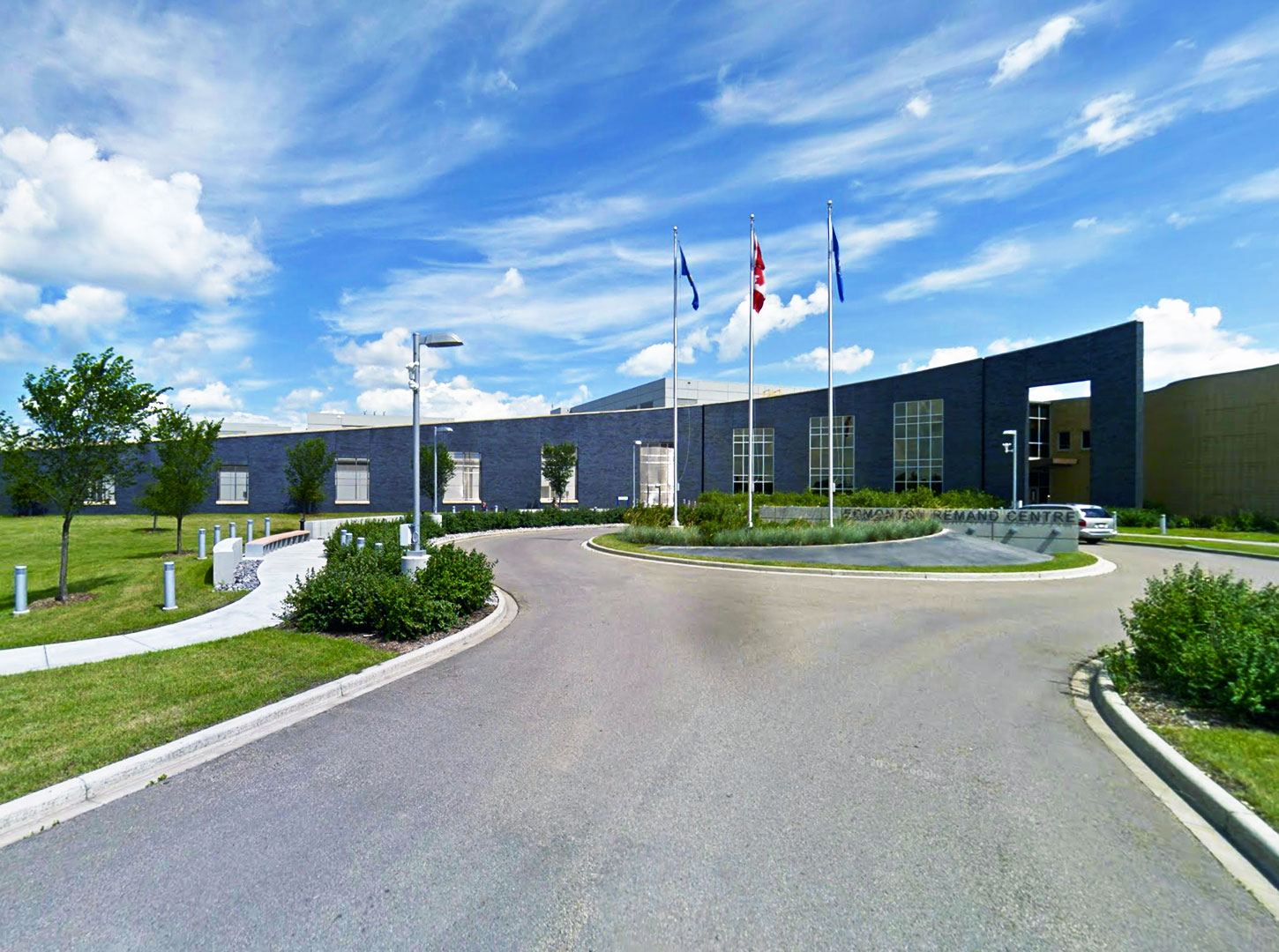 Edmonton Remand Centre – Main Entrance View Street-level view of the Edmonton Remand Centre showing a curved facade, large windows, flagpoles, and a landscaped circular driveway under a partly cloudy sky.