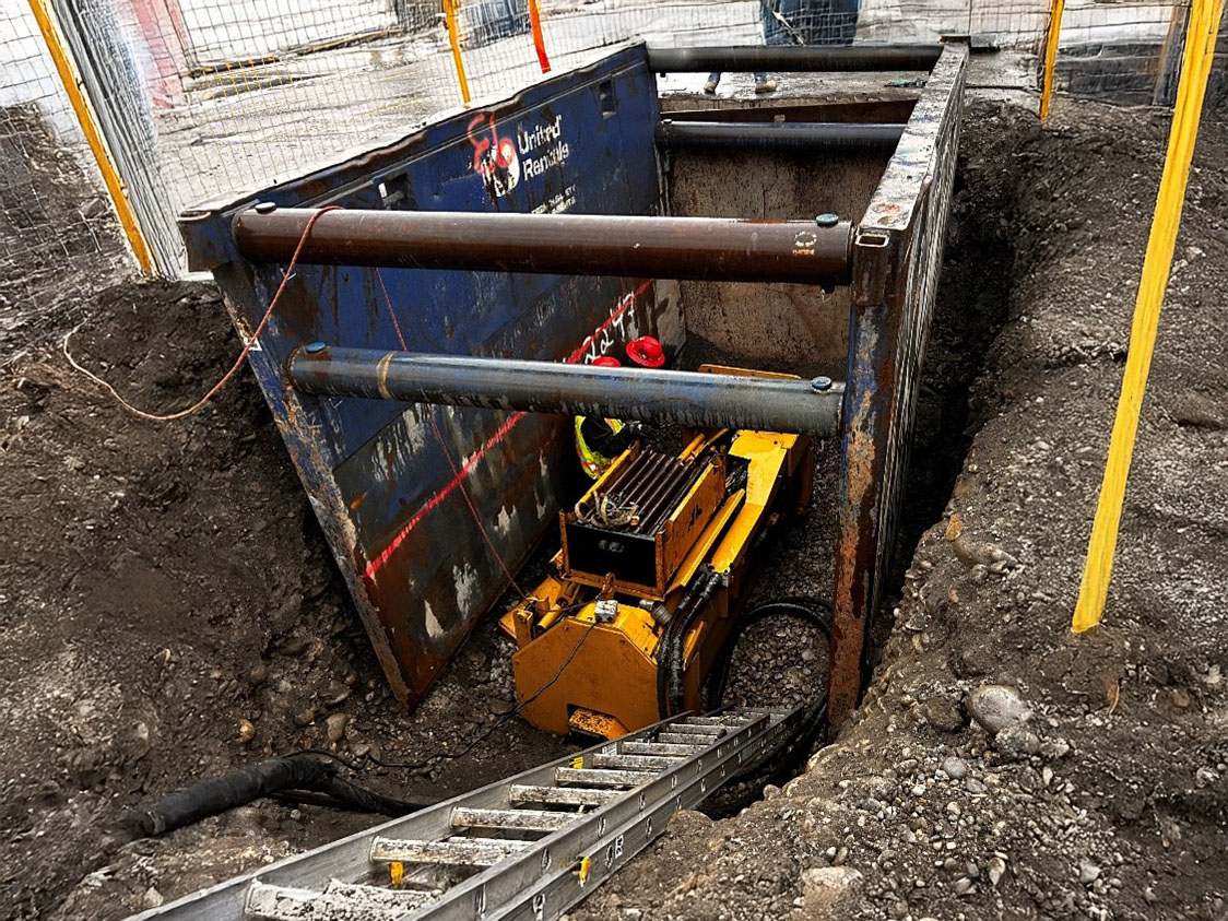 A yellow excavator machine in a reinforced trench at a construction site.