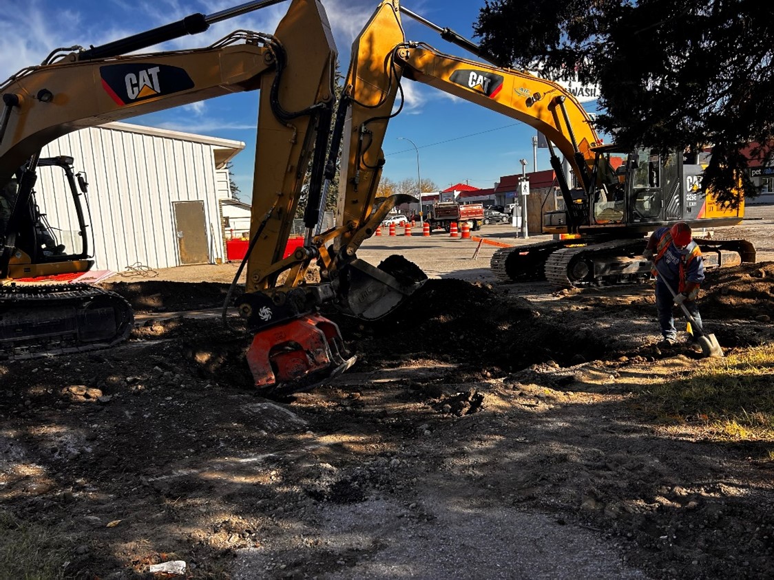 Two track hoes engage in earth moving. A worker in PPE works a shovel nearby.