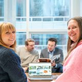 Two individuals out of a gathering of four around a computer in a boardroom look over their shoulders at the camera and smile.