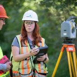 Two individuals in hardhats and high visibility vests are in discussion near a survey total station.