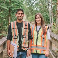 Two people in high visibility vests are standing on a wooden bridge in a forested area, and are smiling at the camera.