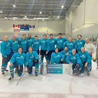 A hockey team in matching teal jerseys stands and kneels around a sign announcing a charity hockey tournament hosted by McElhanney, raising funds for Ronald McDonald House.