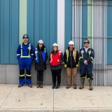 A smiling row of individuals in high visibility outerwear and hardhats stands on a concrete slab in front of a striped teal an blue wall.